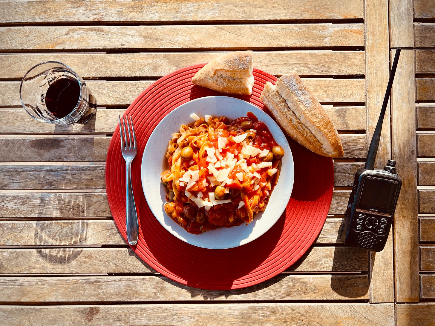 A glass of wine sits next to a plate of pasta, next to some bread and a radio. Long shadows are cast at the end of a warm Spanish day in February 