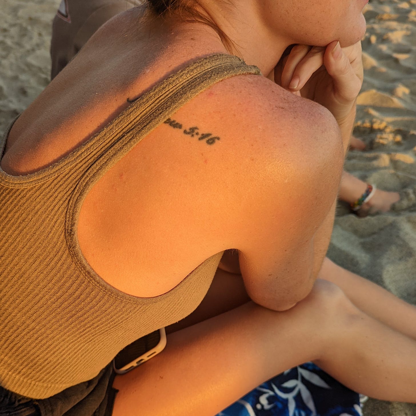 A female figure sits on the sand watching the sunset. She is wearing a tan tank top and on her right shoulder is a visible tattoo which reads Matthew 5:16