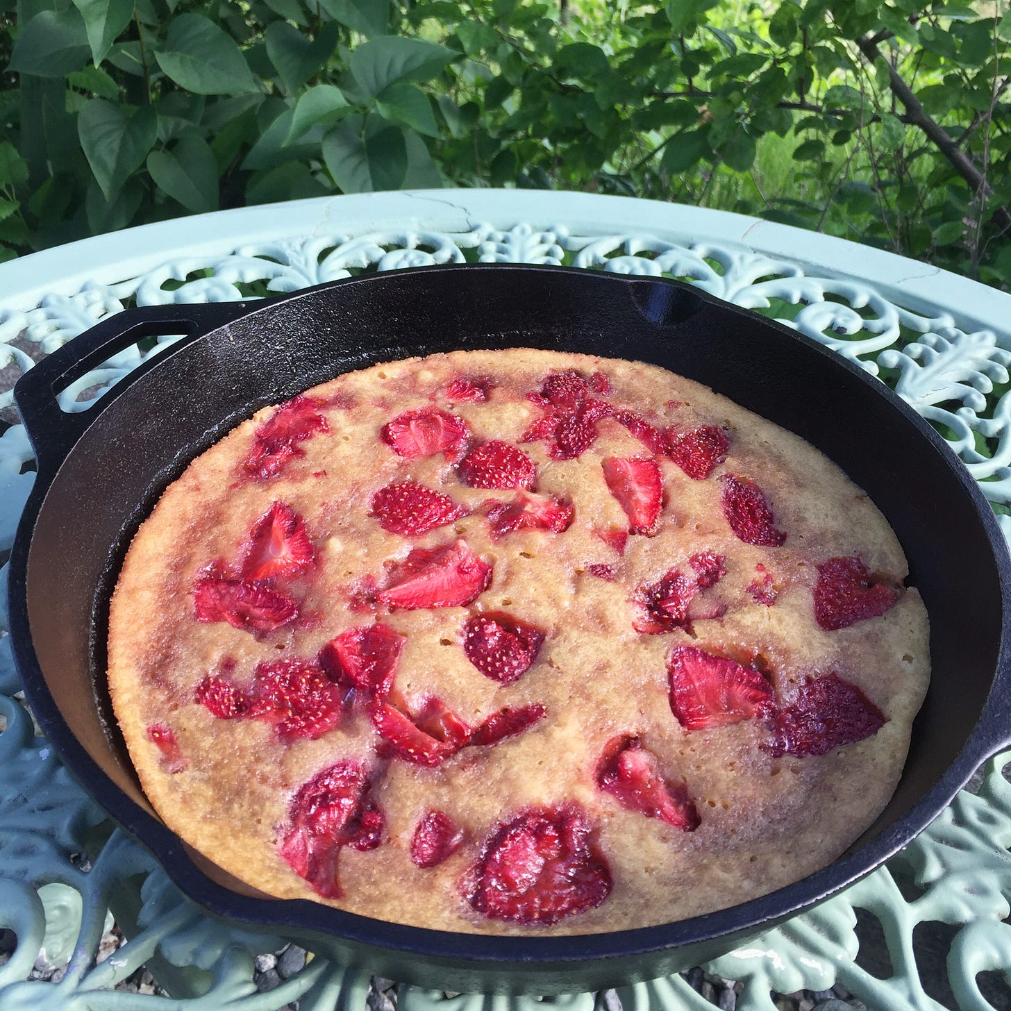 a cast iron pan on top of a wrough-iron table. In the pan is a golden cake with pieces of strawberry sunk into the top.