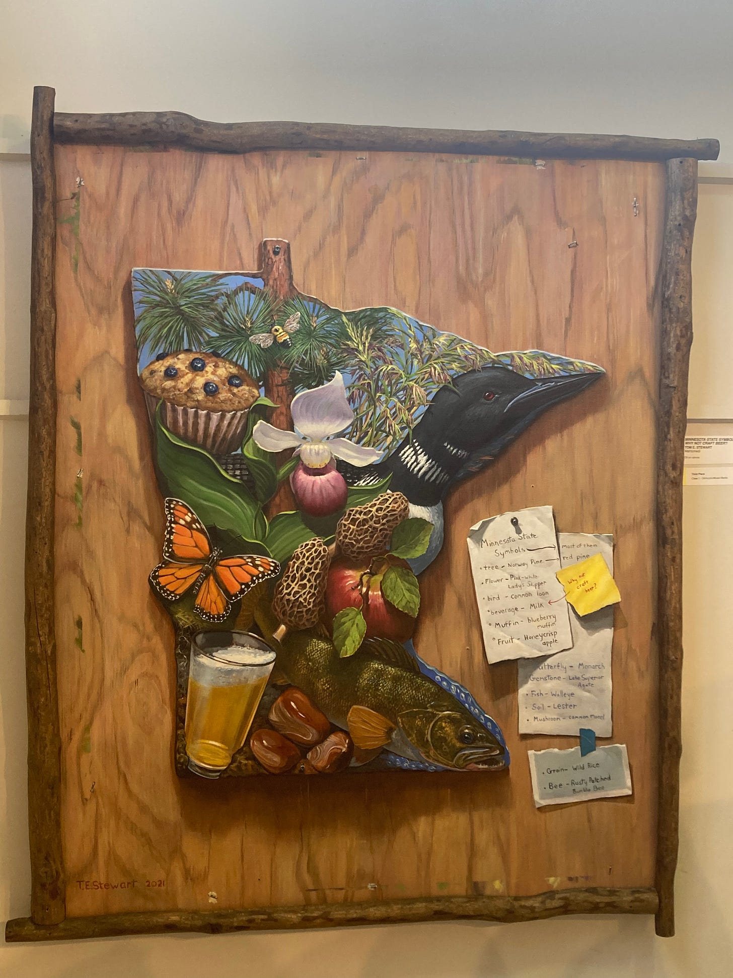 a shape of Minnesota on a wooden background is filled with realistic interpretations of the state's symbols: on the top left, a blueberry muffin, a bumblebee, a pine tree. in the center, a monarch butterfly, a lady slipper flower, mushroom morels. on the top right, wild rice and a loon. in the middle right, an apple. on the bottom right, a walleye. on the bottom left, a glass of beer and some stones.