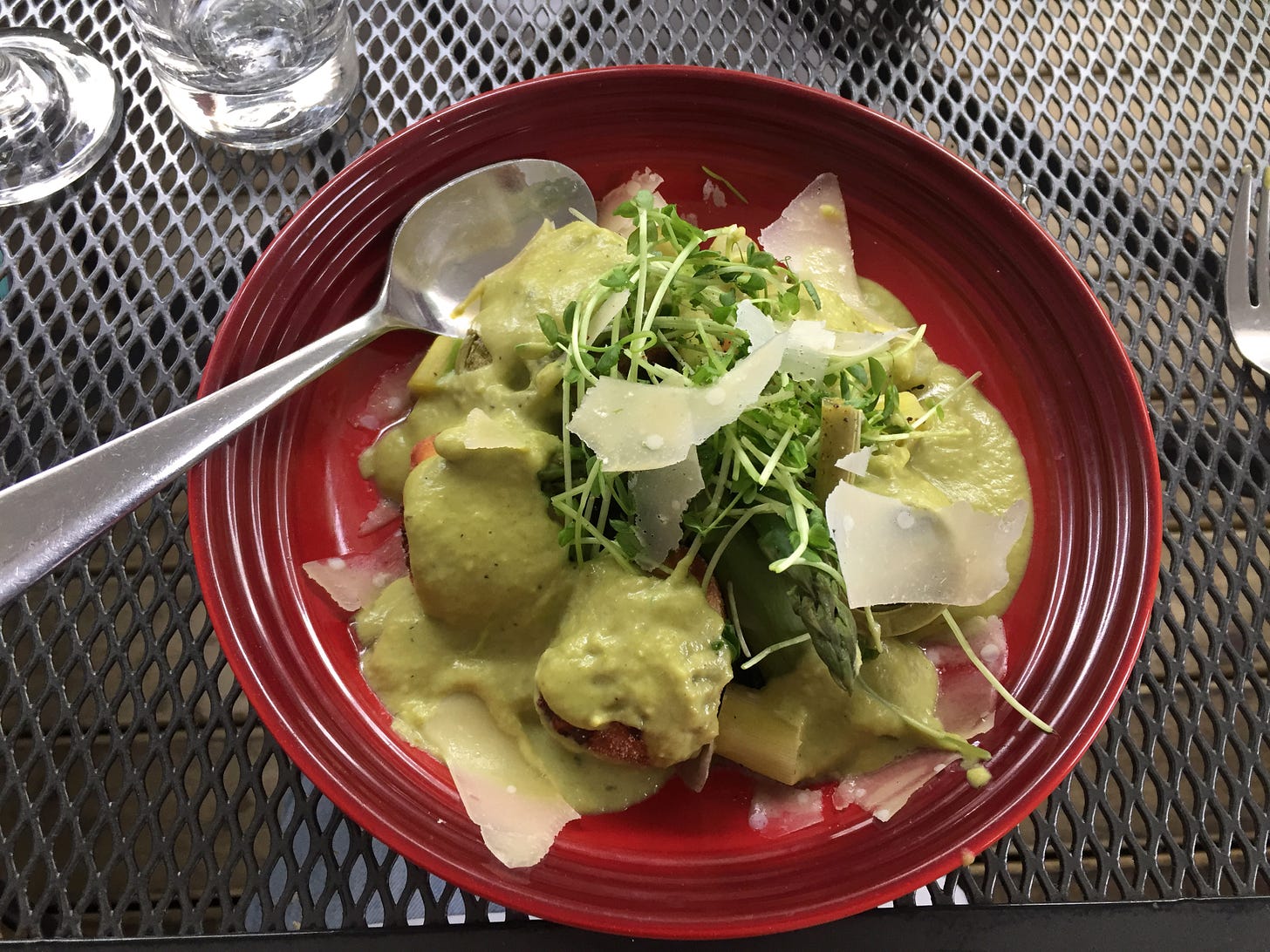 A red bowl of large gnocchi in a light green sauce, with pea shoots, shaved parmesan, and pieces of asparagus. 