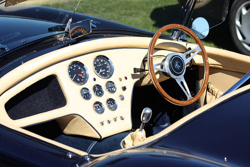 Imagem mostra painel de um carro, com volante, câmbio e vários marcadores similares a relógios, como conta-giros, velocímetro e outros instrumentos.