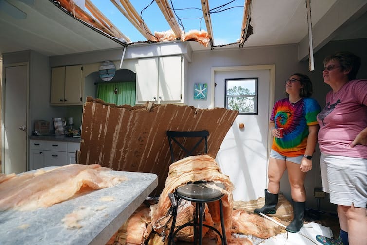 Two women stand in a wind-damaged kitchen looking up at the sky through a missing section of roof.
