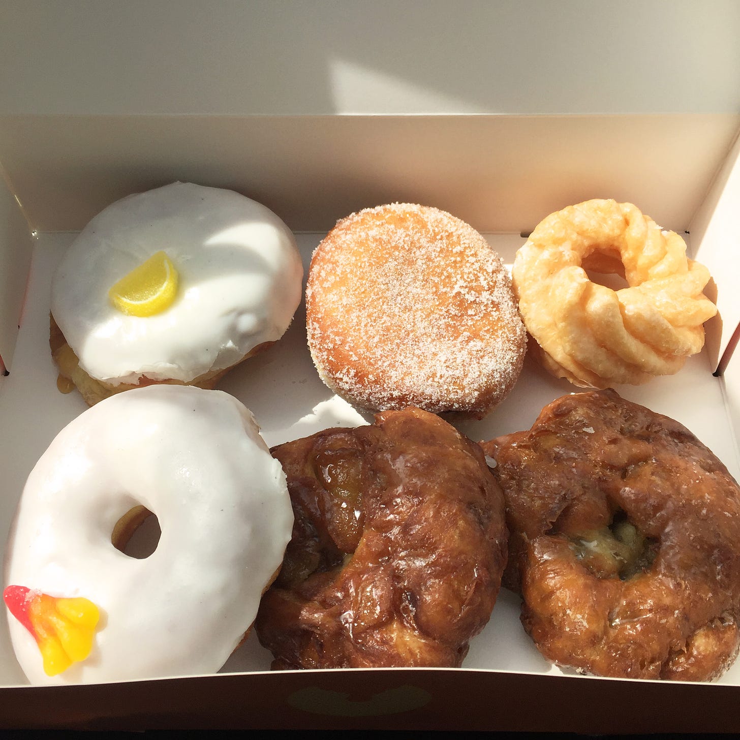 An open box with a white interior, filled with six donuts. Two fritters are in the bottom right, and above them are a cruller and a sugary jam donut, and on the left are two donuts with white glaze, each with a small gummy candy on top.
