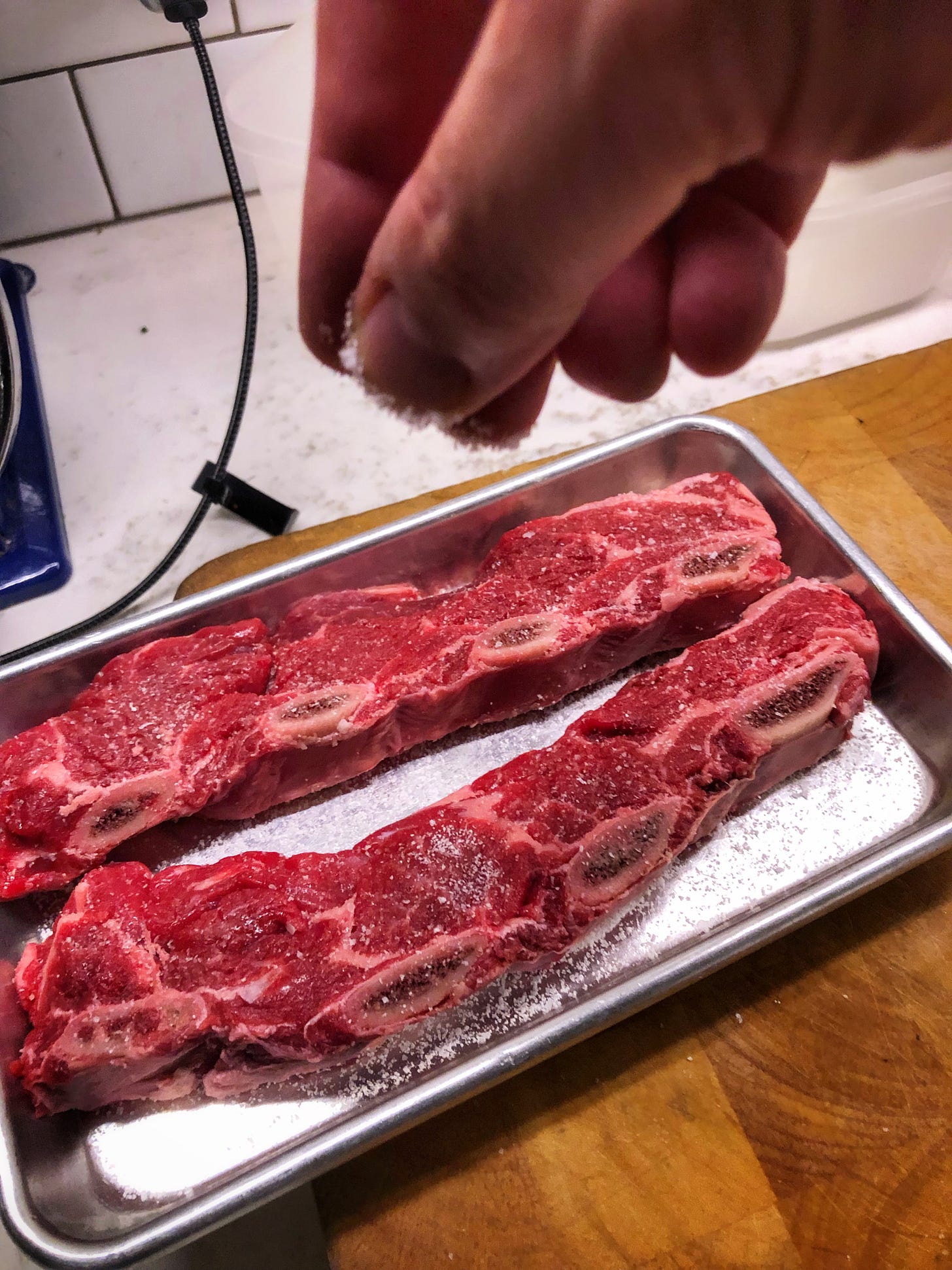 The short ribs are not on the salted sheet tray. The hand sprinkles salt onto the surface of the meat.
