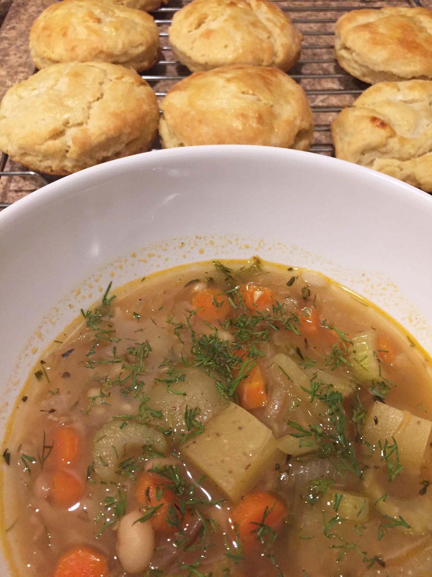 In the foreground, a white bowl of stew with carrots, beans, potatoes, and fresh dill. Behind it, a cooling rack with six browned buttermilk biscuits.