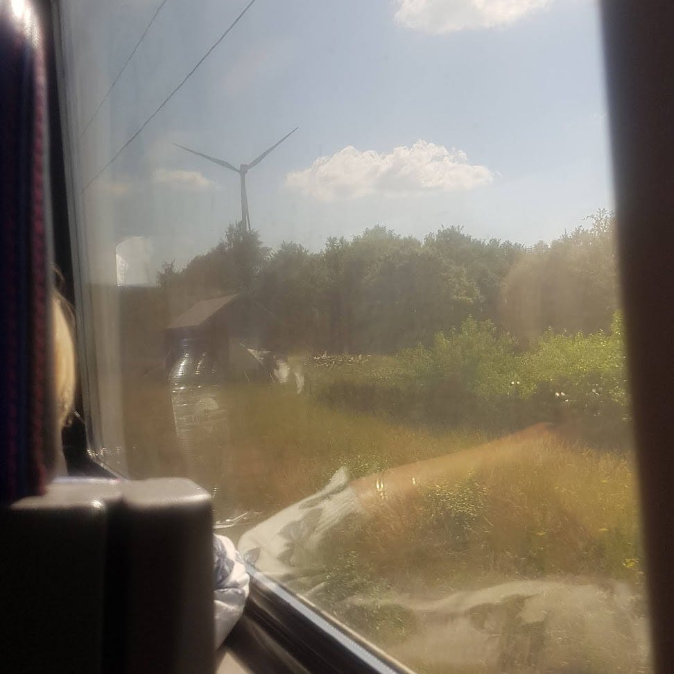 View from inside a train carriage, of a green field and blurry building. A wind turbine is in the distance.