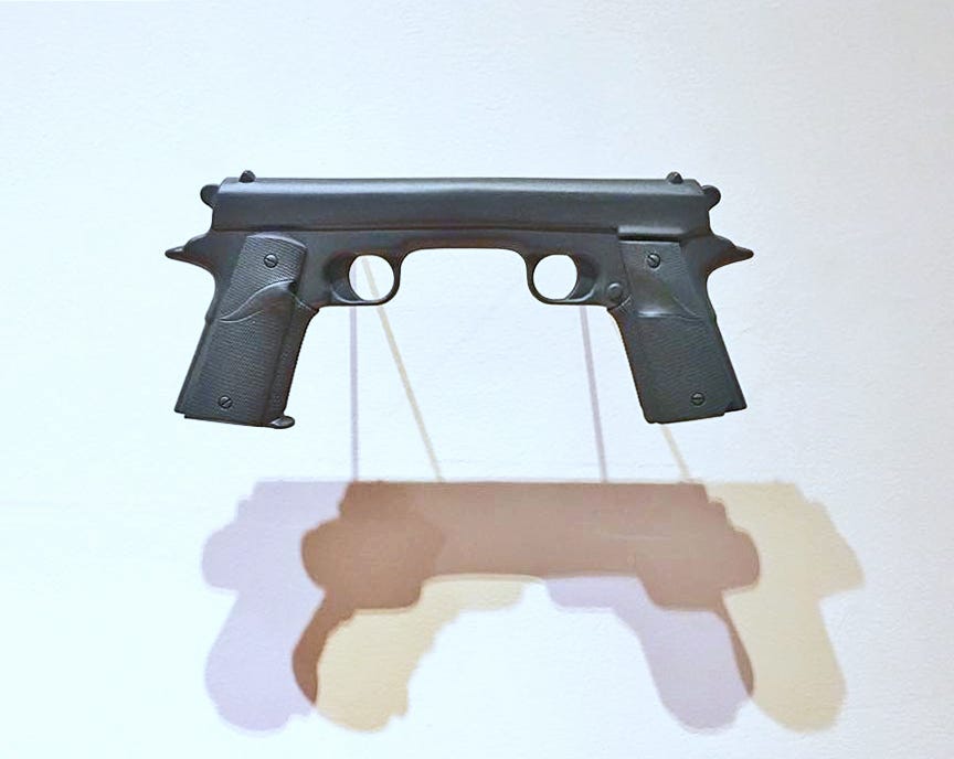 Against a light-blue wall hangs a sculpture of two handguns facing each other. Their barrels have been joined so that the two guns together form one seamless object with two grips, two triggers and one barrel. The light shining on the sculpture casts a stark shadow of the piece onto the wall.