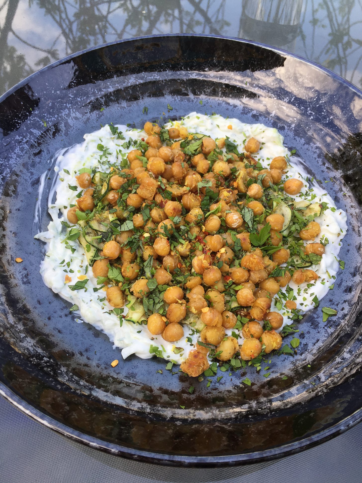 On a glass patio table, a shallow black speckled dish with tzatziki spread in the centre. On top of the dip is sautéed slice of zucchini covered with crispy chickpeas, dusted with herbs and chili flakes.