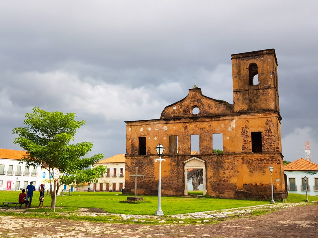 Foto colorida da praça central de Âlcantara, com as ruídas de uma igreja no meio de um gramado verde e casas históricas em volta