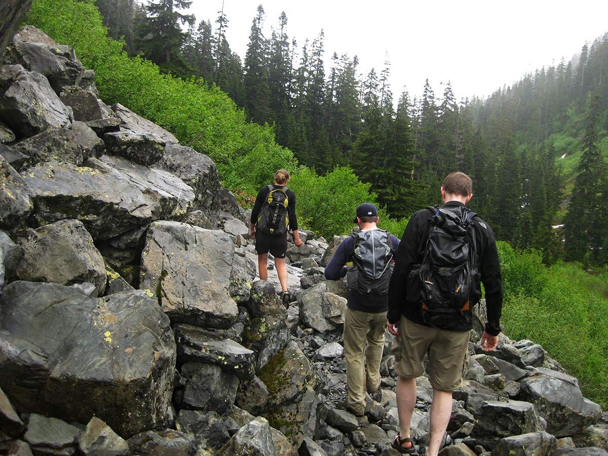 three hikers cross a talus field on the Denny Creek / Melakwa Lake trail, brushy green willows and dark conifers ahead