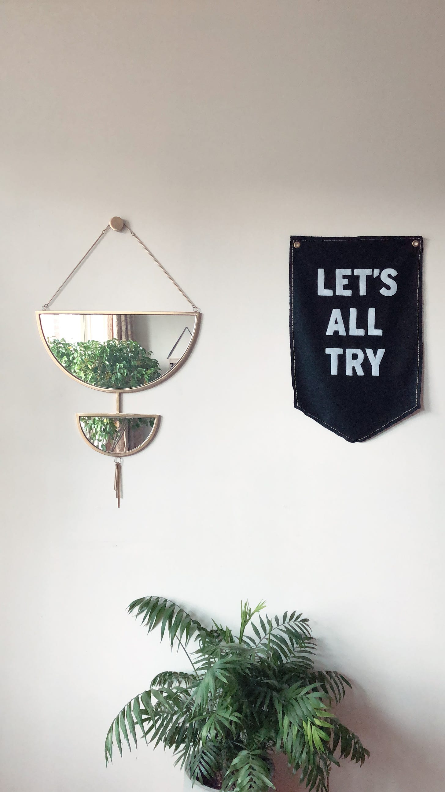 A geometric mirror hangs above a house plant and beside a black and white banner that reads "Let's all try."