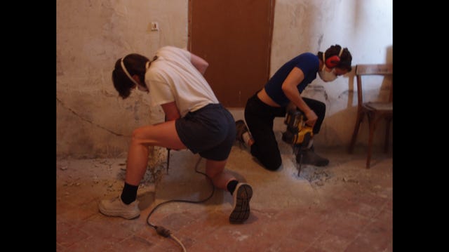 Two dancers are using jackhammers to demolish an old concrete ramp. They are in the same position but mirroring each other, in a kind of satisfying symmetry. They have on protective masks, goggles, and earplugs.