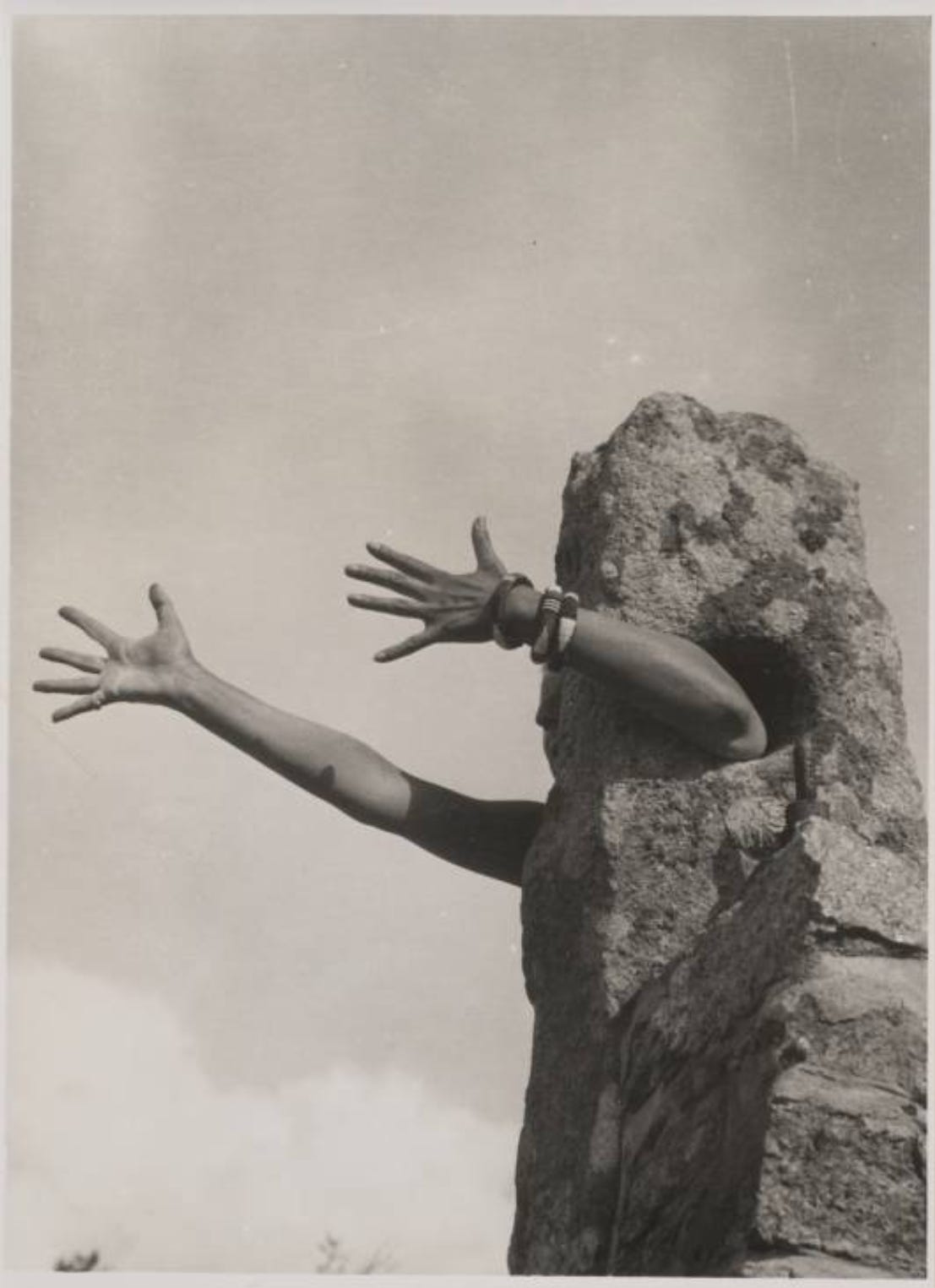 A self-portrait by Claude Cahun. Their arm is stuck through a hole in a person sized rock so they look like they're a rock with arms, extending their arms forward as if to hug somone.