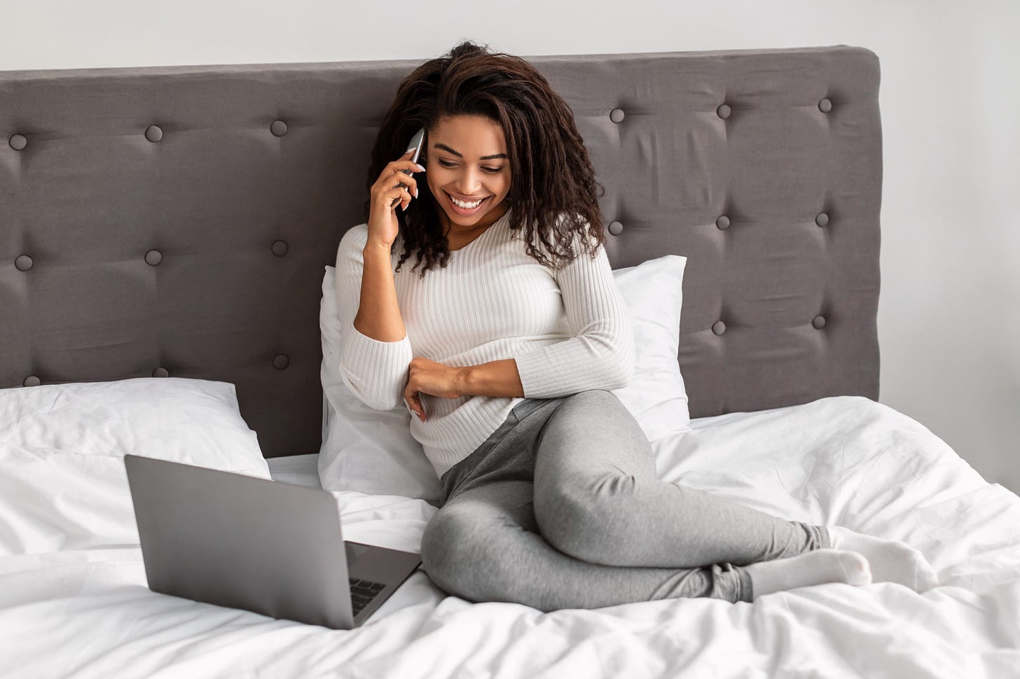 A happy Black woman sits on the bed with her laptop beside her. She smiles as she chats on the phone.