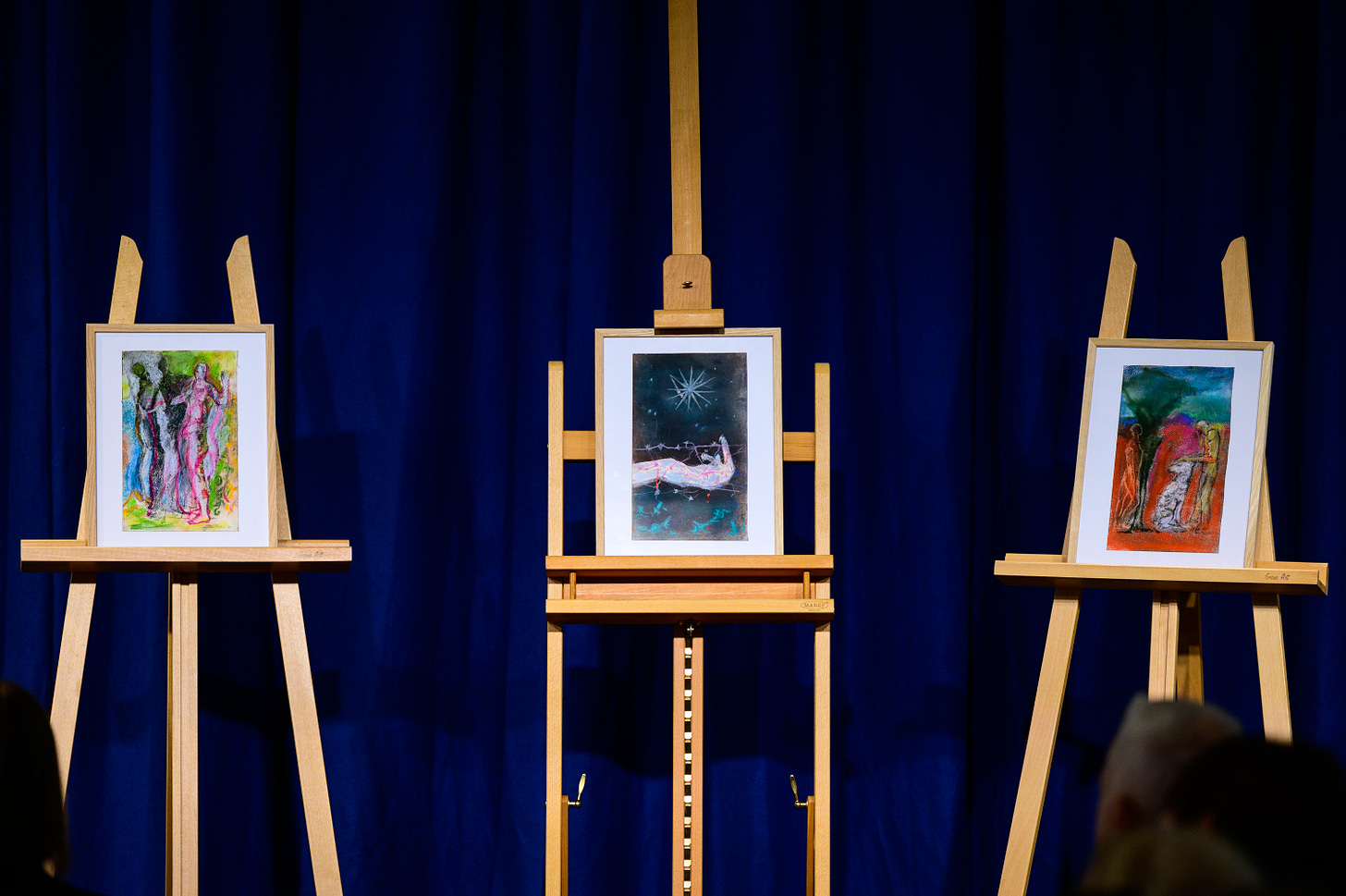 Three art pieces on wooden easels in front of a blue curtain. 