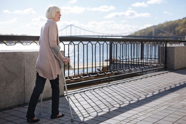 Tall, gray-haired white women walks with Canadian crutches along a river walkway, which has an ornate grating along her left side. The grating casts a striking shadow on the sidewalk in front of her. 