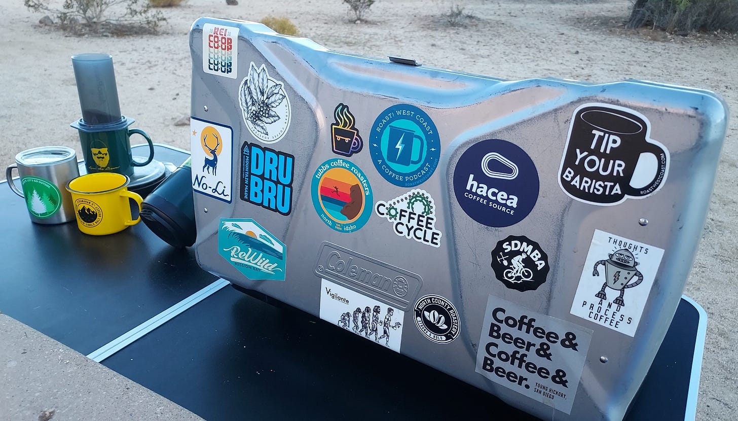 The lid of a silver Coleman propane camp stove is covered in coffee shop stickers and sits on a table with several coffee mugs and an Aeropress brewer. The photo is taken at an angle viewing down and the ground in the background is desert sand.
