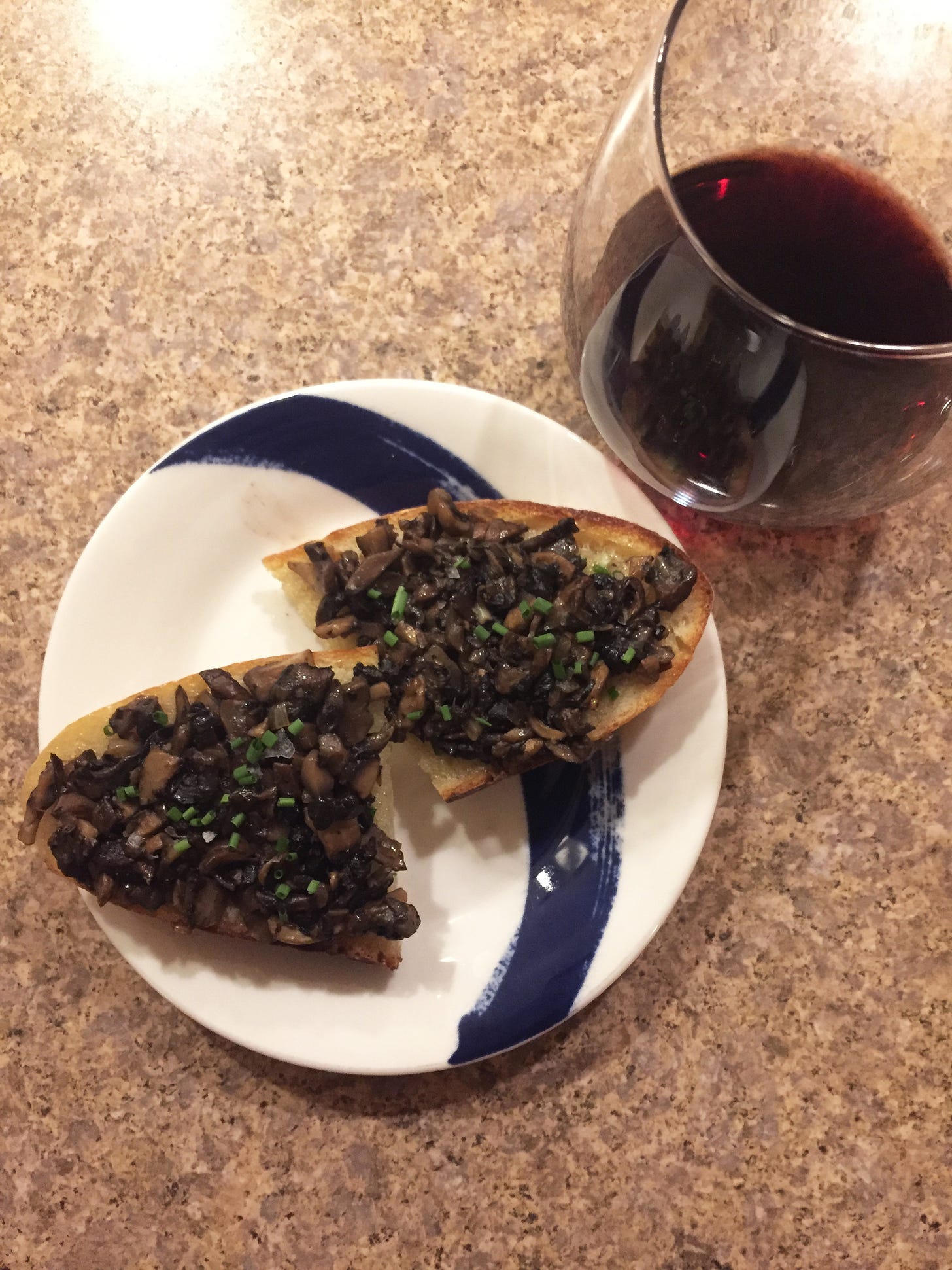 Two halves of a large slice of sourdough, each topped with the chopped fried mushrooms described above, and sprinkled with chives. A stemless glass of red wine sits at the upper right edge of the plate.