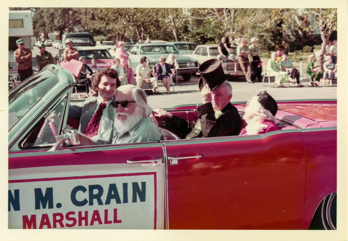A color photo of a red convertible in a small-town parade. In the back seat, an old man is tipping his top hat to the camera. His wife, in a black toque, is seated beside him. Spectators at the side of the road are standing and sitting on lawn chairs.