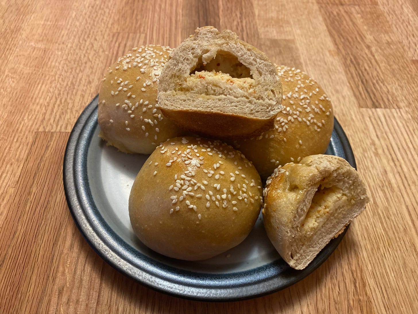A small plate holding three golden brown buns topped with sesame seeds. A fourth bun, piled on top of the others, is cut in half, showing the feta filling.