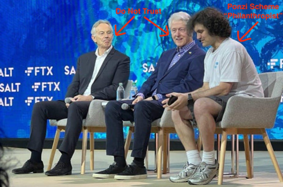 3 white men sit on chairs on a stage. The 2 older white men are dressed in suits and holding microphones. The younger white man is dressed in a tshirt and shorts, wearing a microphone headset. Behind them is a blue background with "FTX" and other letters emblazoned on it in a horizontal pattern. Added by me in red is "Do Not Trust" written with arrows pointing to the heads of the older men (Tony Blair and Bill Clinton), and "Ponzi Scheme Philanthropist" with arrow pointing to 3rd man who is SBF.