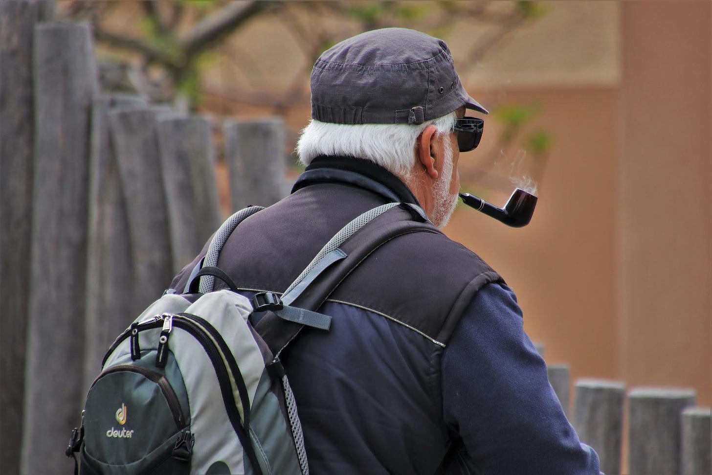 Camera looks over shoulder of older man with gray hair and beard and sunglasses. He is smoking a pipe.