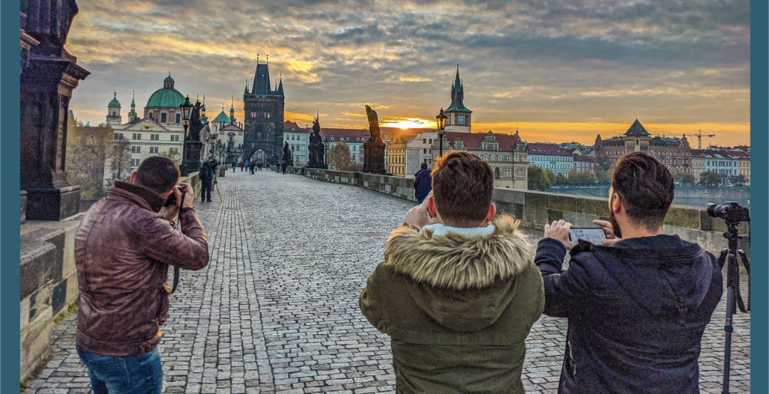 Three photographers standing on the Charles Bridge taking pictures of the sunrise over Prague, Czechia.