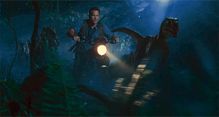 A white man riding a motorcycle through a dark jungle. He is surrounded by a pack of raptors who run alongside him.