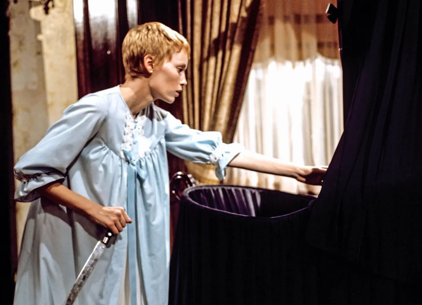 A white woman in a blue nightgown, played by actress Mia Farrow, stands holding a knife over a black crib