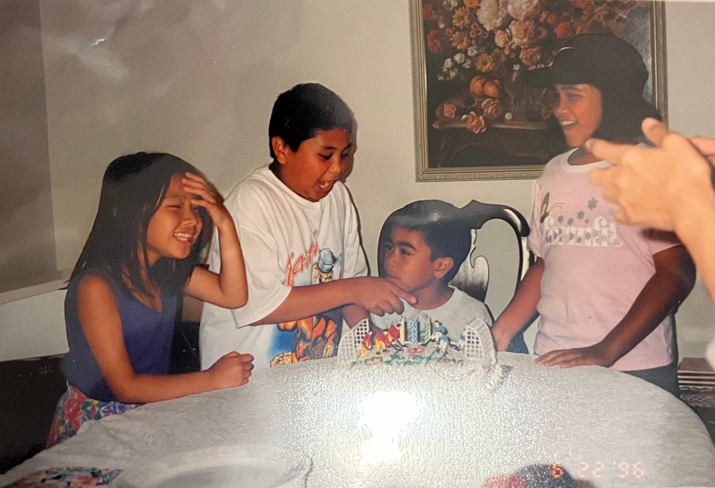 Four children sitting by a dining table. One boy is sitting in front of a birthday cake while another boy is pointing at it. 