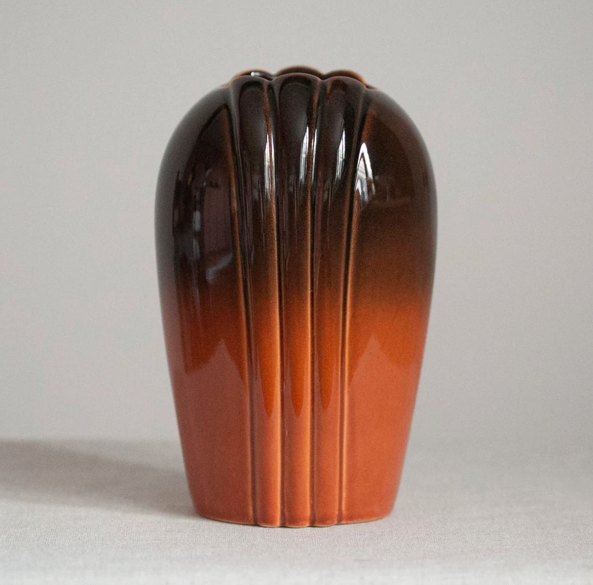 A stunning ceramic vase made to look like a clamshell with three narrow ridges on each side. The glaze grows from a deep chocolate brown at the top to a beautiful burnt orange near the bottom of the vase.
