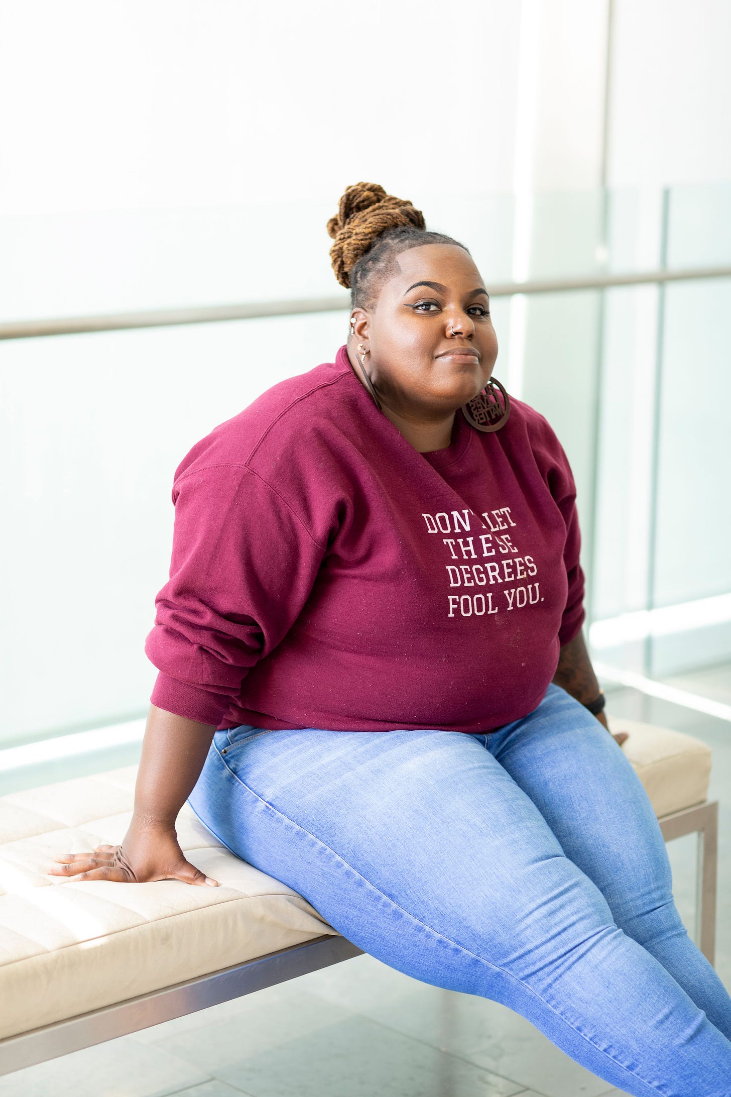 Headshot of Tamice Spencer sitting on a beige leather bench wearing a maroon sweatshirt that reads, “Don’t let these degrees fool you,” paired with light wash blue jeans. 
