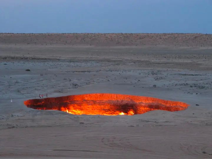 The photo is of a desert with a huge hole pit in the ground lit up red with fire, some flames can be seen licking up, and some people can just barely be seen at one edge, the hole is so big