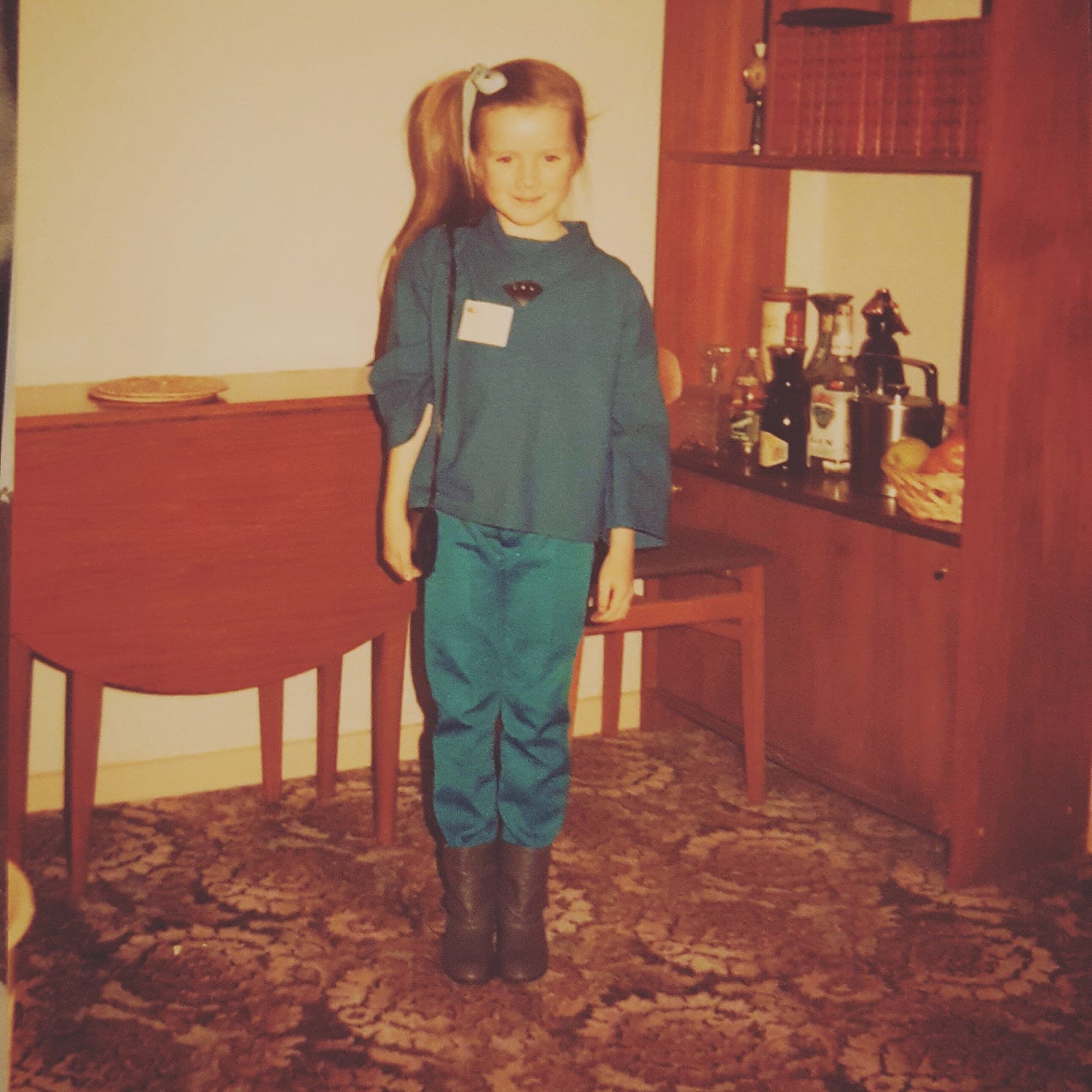 Photo from the 1980’s of a young girl wearing blue trousers and a blue top and grey boots. She has a handbag over her right shoulder and is stood on brown patterned carpet in a room with a brown table and a brown cabinet with bottles of alcohol on it. Photo from the 1980’s of a young girl wearing blue trousers and a blue top and grey boots. She has a handbag over her right shoulder and is stood on brown patterned carpet in a room with a brown table and a brown cabinet with bottles of alcohol on it.
