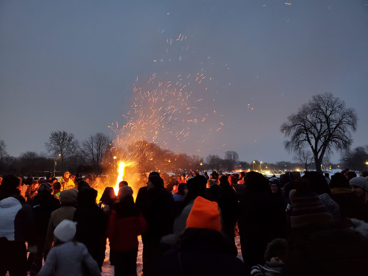 A large group of people stands around a roaring bonfire in the middle of a snowy park.