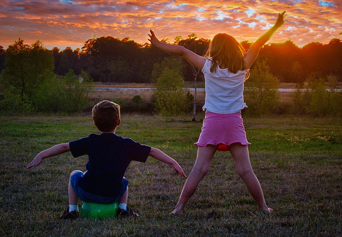 two children facing a colorful sunset on the lake, a boy sitting on a green ball arms outstretched and the girl in a white shirt and pink skirt standing with feet and arms stretched out