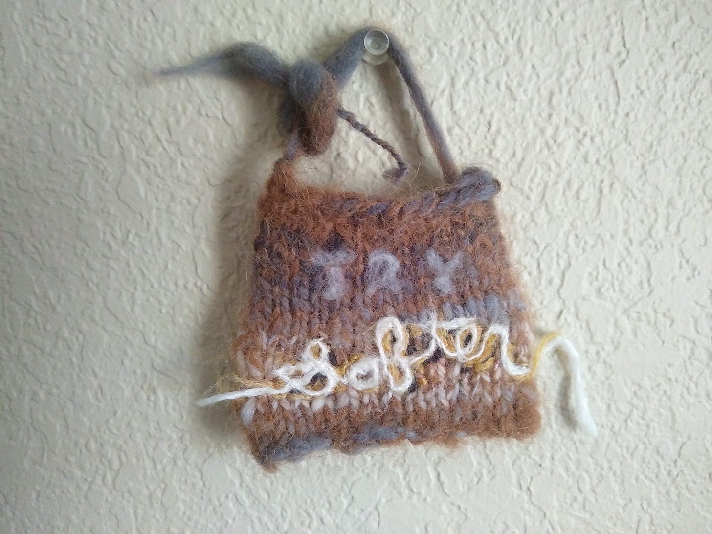 A brown and grey knitted swatch hanging from a thumbtack, with Try Softer felted onto it. The swatch.