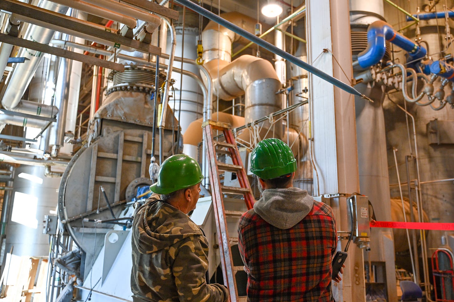 two men seen from behind in an industrial setting looking at a large piece of equipment. both men wear warm layers and hard hats. the man on the left observes and looks ahead while the man on the right points towards the equipment as if explaining its operation.