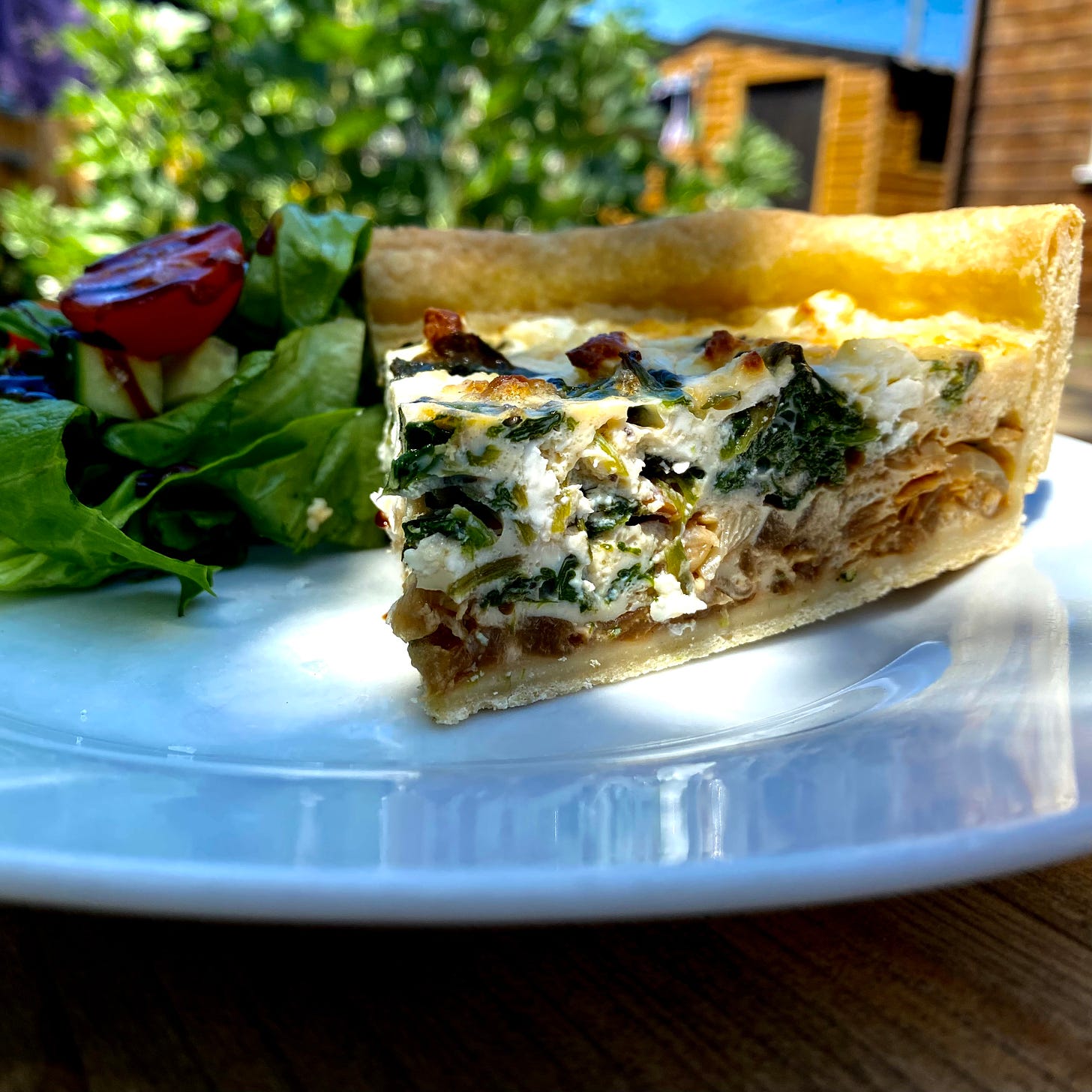 close-up picture of a spinach and cheese tart, side salad to the side on a white plate. Wooden buildings and foliage in the background.