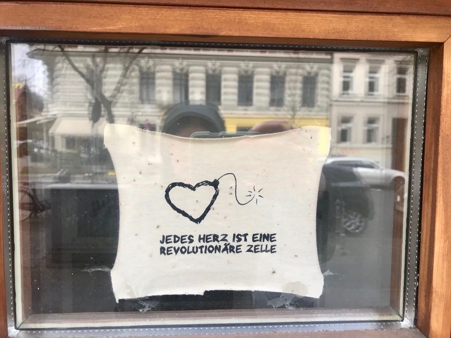 An einer Fensterscheibe klebt ein Stück Stoff mit dem Spruch "jedes Herz ist eine revolutionäre Zelle." In der Scheibe spiegeln sich die Straße, Autos, ein Haus und die fotografierende Person mit ihrem Handy.