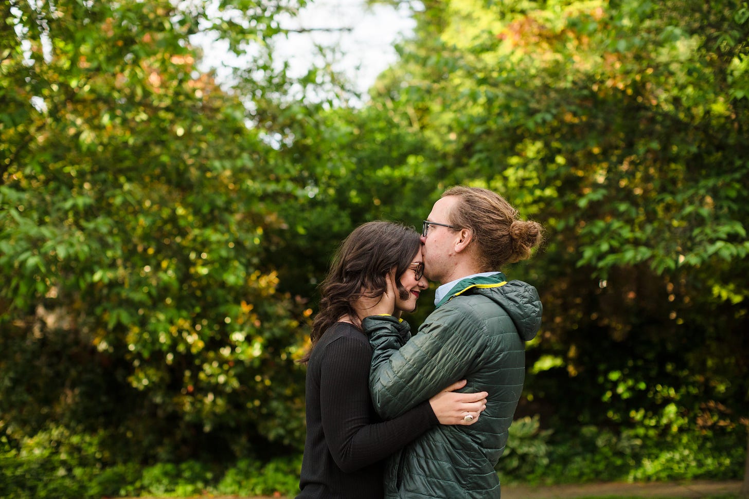 Travis and Kaitlin are standing outside in front of some trees. Their arms are around each other, and Travis is kissing Kaitlin's forehead, and she is smiling.