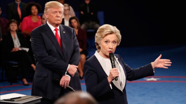 Hillary Clinton standing on stage speaking into a mic while donald trump stands behind her scowling