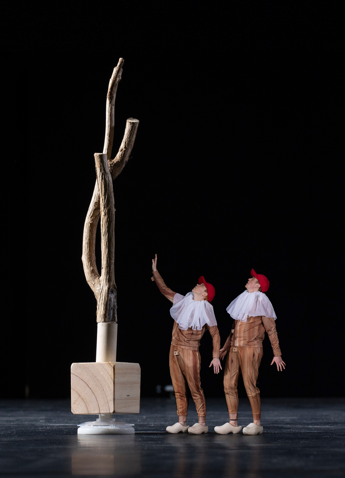  Artists Cade &amp; MacAskill are wearing wood patterned leisure suits with white clown-like ruffs and red baseball caps. They are looking up at a giant twig which looks like a tree because of forced perspective in the photo.  