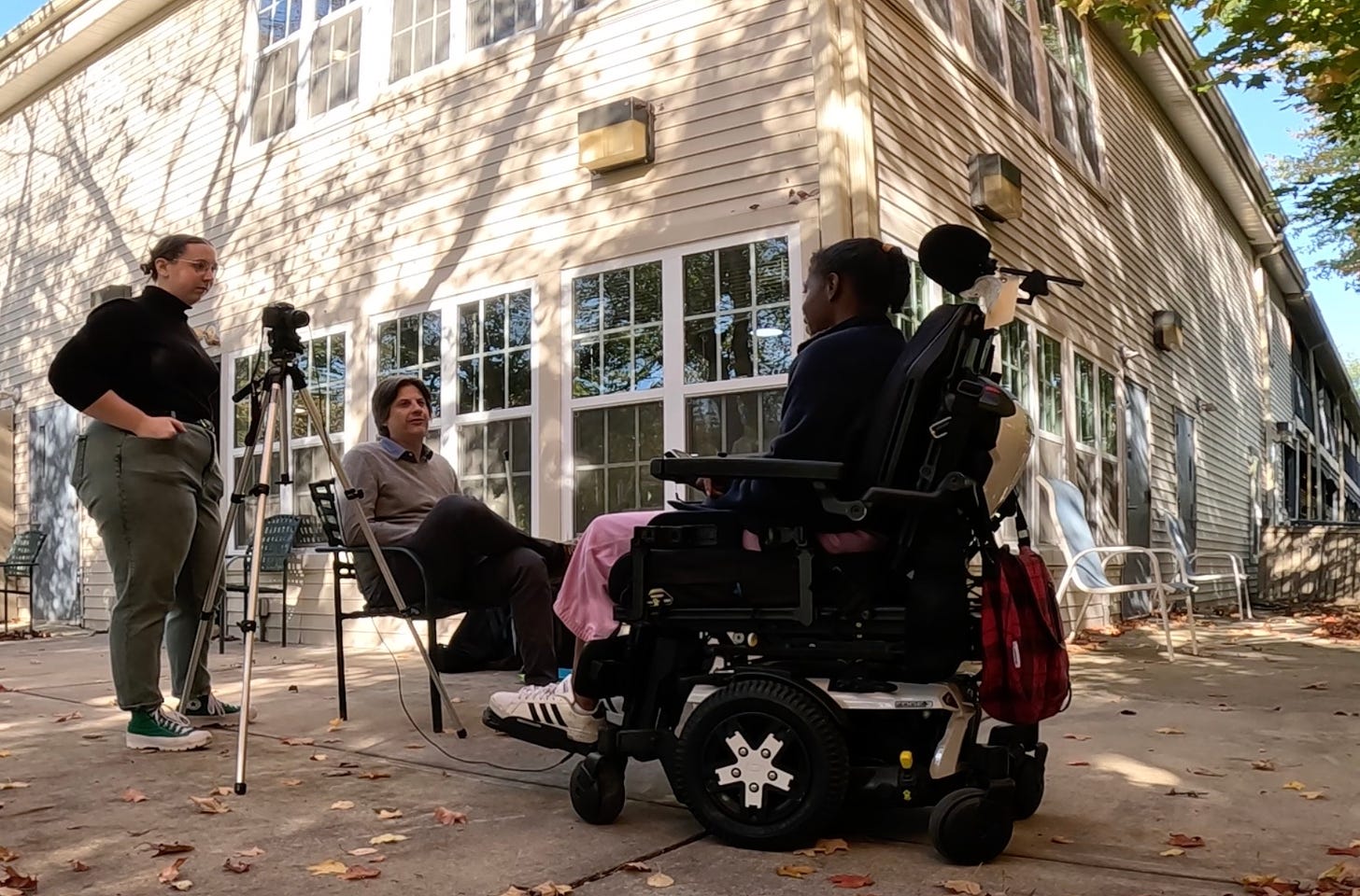  A woman in a power wheelchair is seated at an outdoor patio. Ahead of her is a man seated in a chair and a woman standing behind a camera that is mounted to a tripod. 