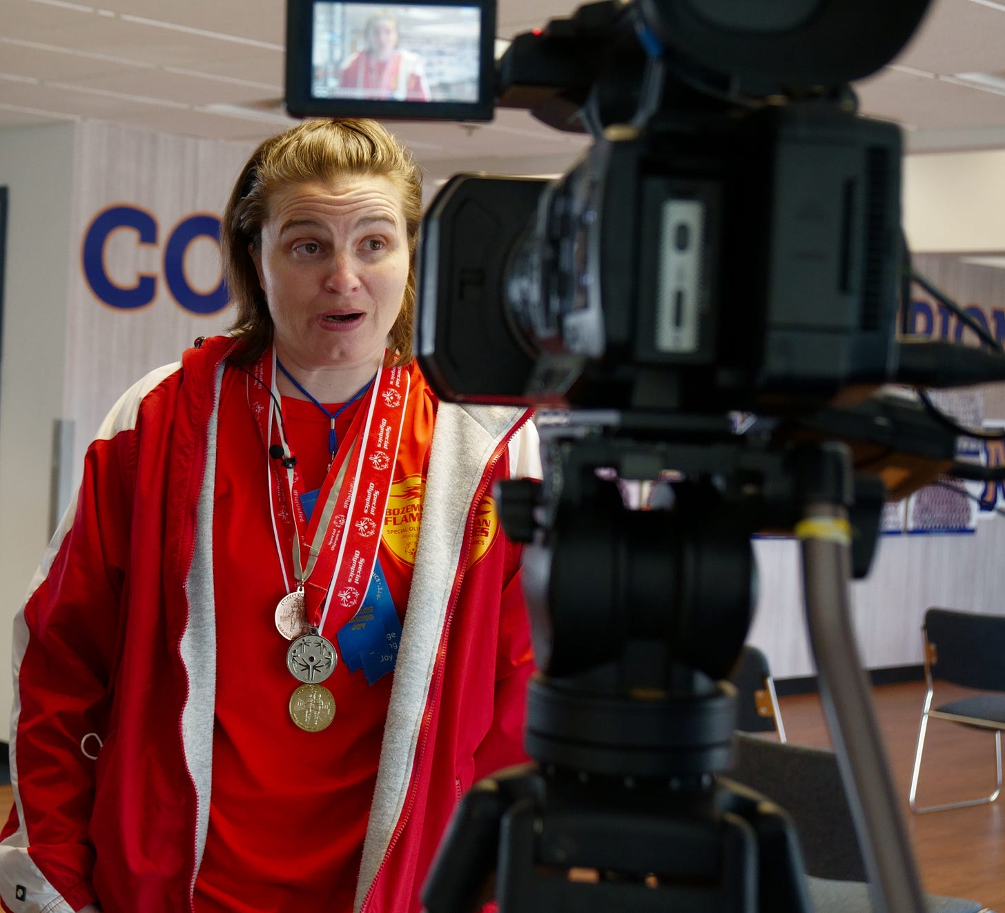 A young woman with short light brown hair and wearing her red Bozeman Flames team apparel speaks into a TV camera. She is wearing several Special Olympics medals and blue ribbons. Her image is visible in the monitor of the television camera in front of her. A young woman with short light brown hair and wearing her red Bozeman Flames team apparel speaks into a TV camera. She is wearing several Special Olympics medals and blue ribbons. Her image is visible in the monitor of the television camera in front of her.
