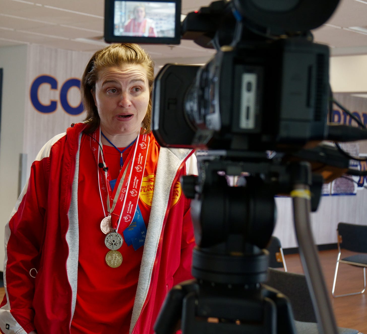 A young woman with short light brown hair and wearing her red Bozeman Flames team apparel speaks into a TV camera. She is wearing several Special Olympics medals and blue ribbons. Her image is visible in the monitor of the television camera in front of her.