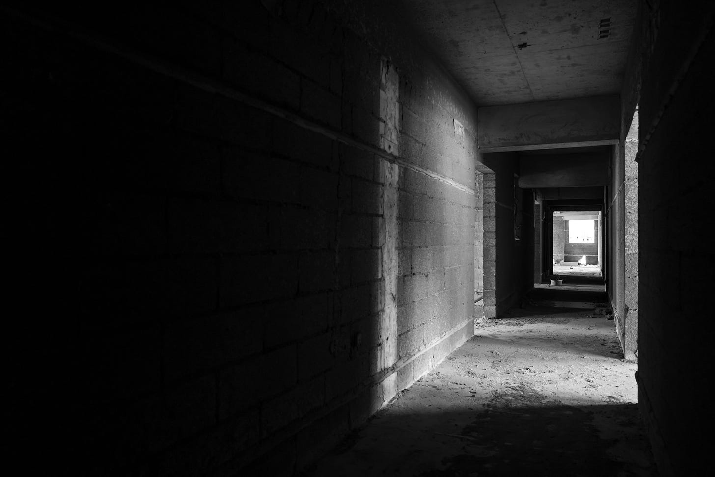 A black and white image of a hallway in an abandoned building. The walls are made of concrete blocks, and the floor surface is peeling. There are shadows and bands of light along the length of the hallway.