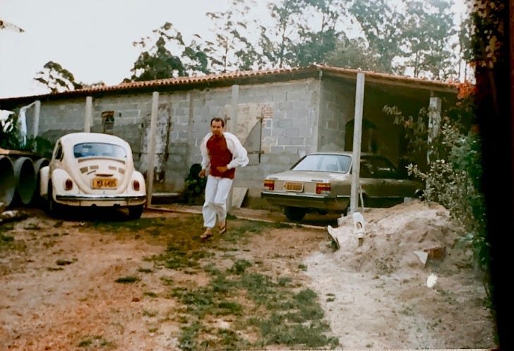 foto do meu pai, um homem pardo vestindo camiseta bordô e conjunto de moletom cinza. ele corre de chinelo de dedo em direção à pessoa que tira a foto. do seu lado direito há um fusca bege. do lado esquerdo há um chevette cinza. o chão é de terra batida e ao fundo dá pra ver uma casa com tijolos aparentes, sem reboco. há um monte de areia em sua frente, para construção da casa. o dia está nublado.