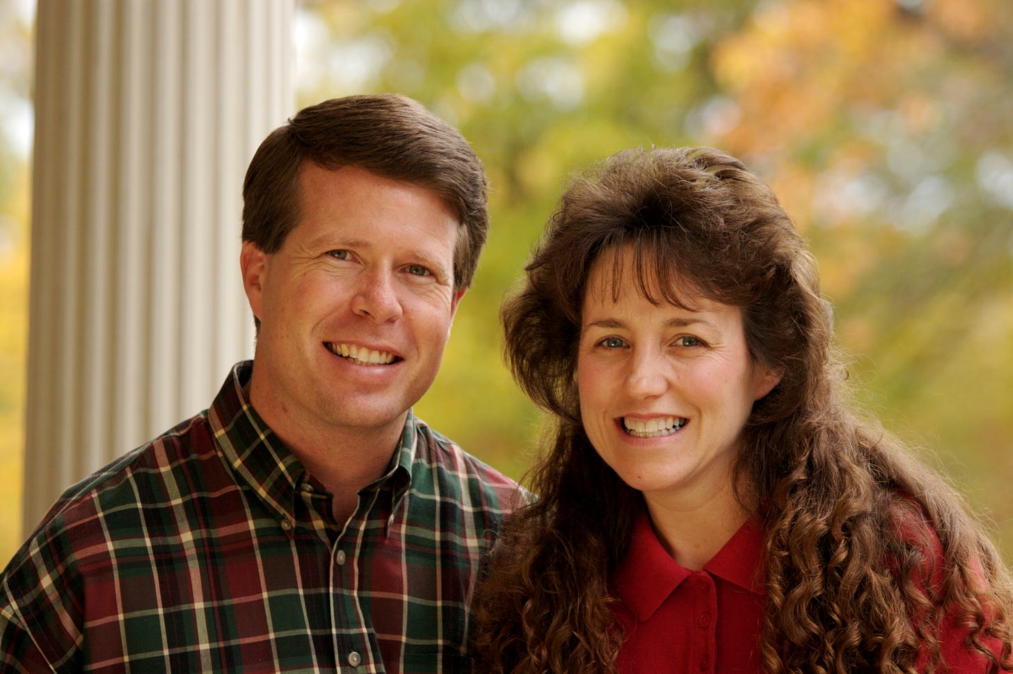This is a wikipedia photo of Jim Bob and Michelle Duggar, probably several years old at this point. They're outside with blurry trees in the background and standing in front of an off-white column of some sort. They're both white people with brown hair in case you didn't know, and they're both smiling with very old-fashioned hair. Michelle is wear some sort of red collared dress and Jim Bob is wearing a green, red, and yellow plaid button-down shirt. they suck